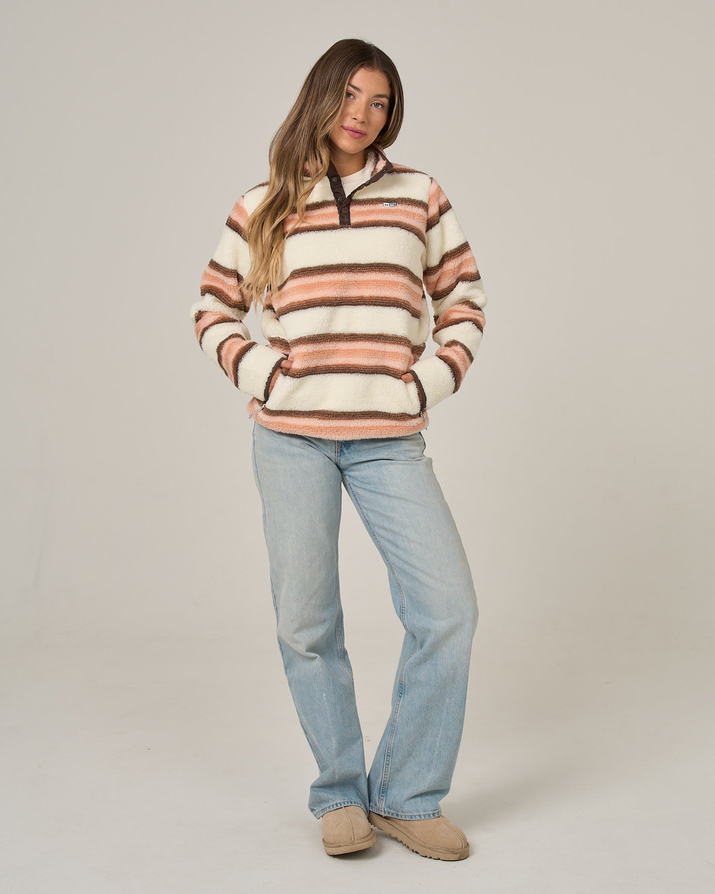 A person stands against a plain light background with hands in the pockets of a Salty Crew Calm Seas Pullover in Dusty Coral, paired with light blue jeans and tan shoes, wearing a relaxed expression.