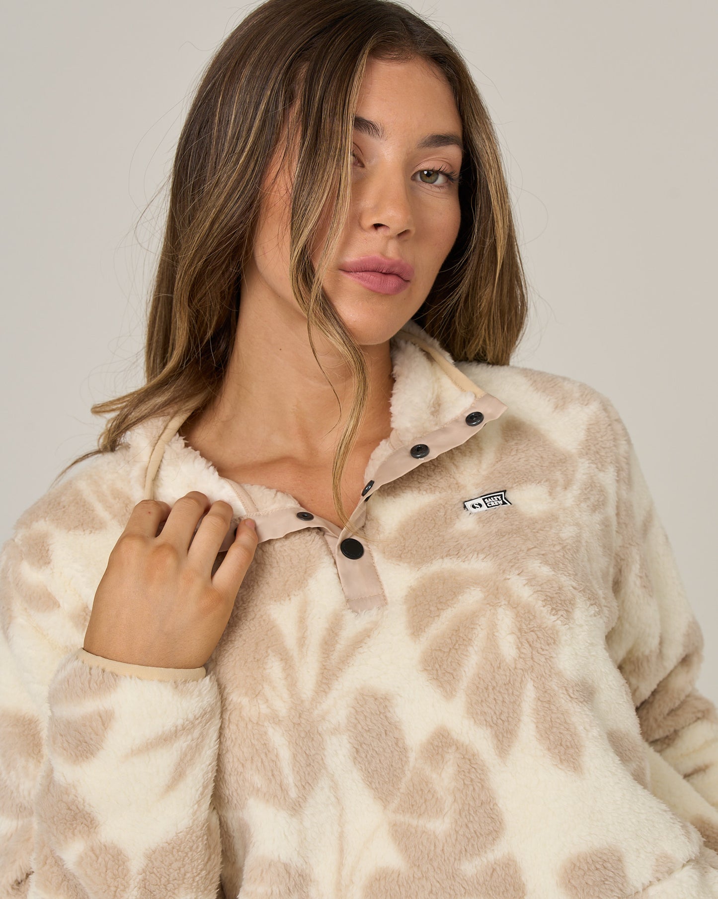 A woman with long hair wears the Salty Crew Calm Seas Pullover in Natural—a cream and beige patterned sherpa fleece featuring a mock neck, black buttons, and a small chest logo—posing with one hand near the collar against a plain background.