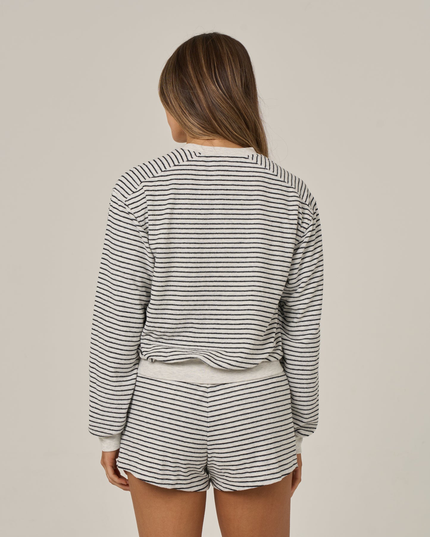 A woman stands with her back to the camera, wearing Salty Crew’s Leeward Crew in Light Heather—a long-sleeved crew neck top and matching shorts. Her brown hair falls loose as she poses against a plain beige background. This set, crafted from a soft cotton-polyester blend, combines comfort with versatile style.