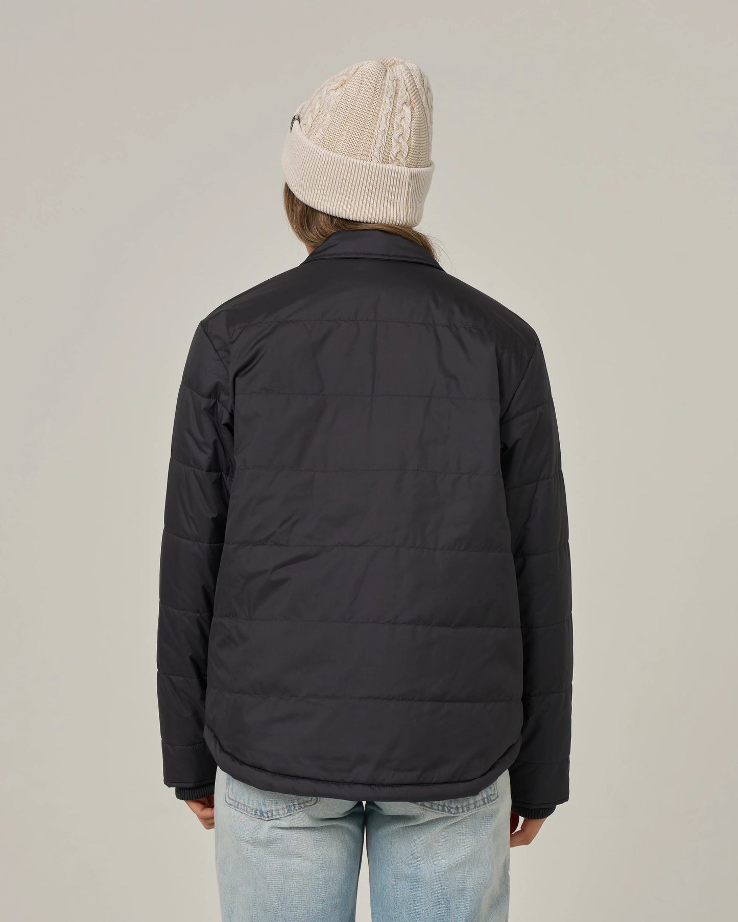 A person facing away, wearing a beige knit beanie, light blue jeans, and the Salty Crew Barrier Snap Jacket in black, stands against a plain light background.