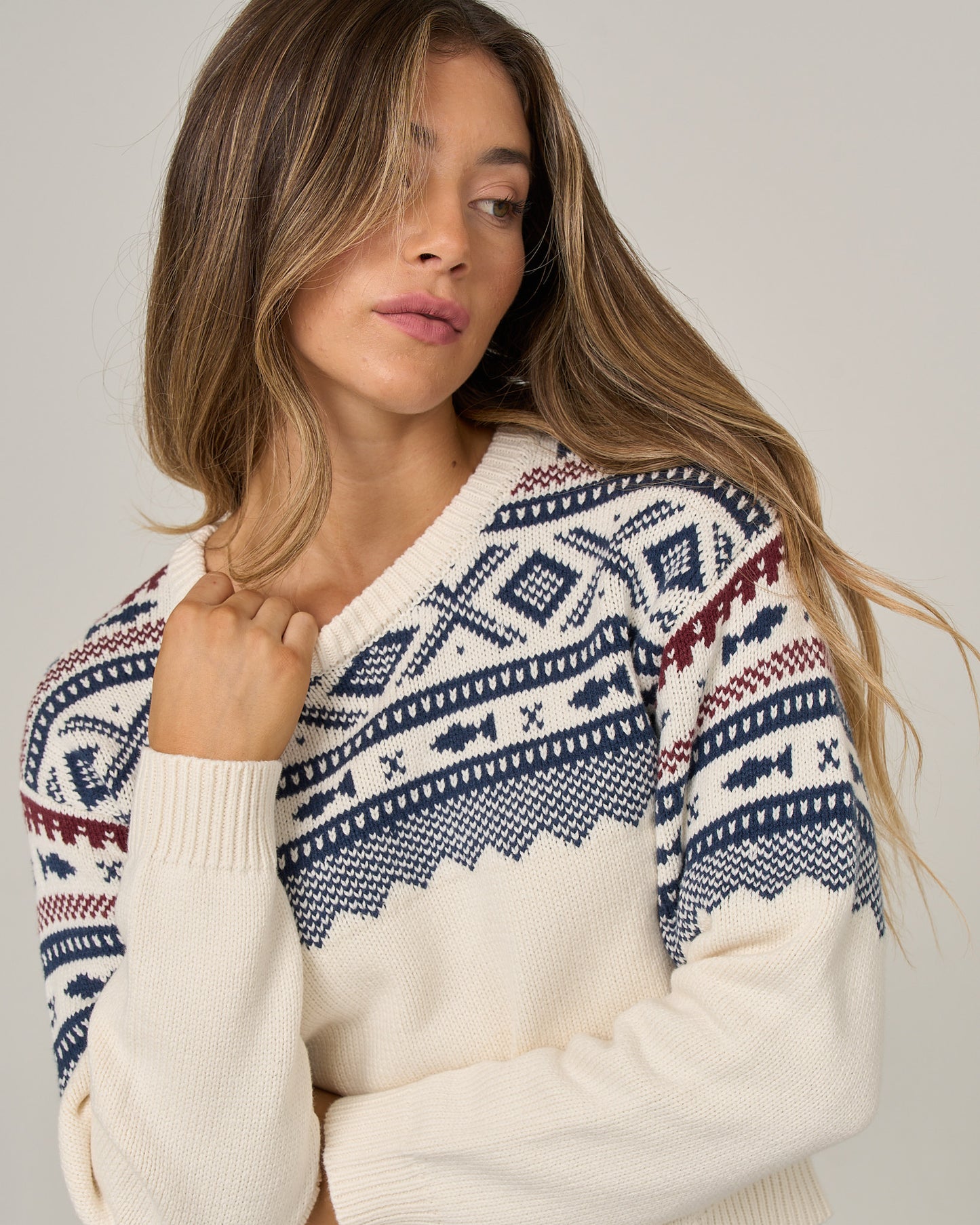 A woman with long, wavy hair wears the Salty Crew Harbor Sweater in Off White, which features blue and red jacquard patterns. She is looking to the side with her hand touching the sweater near her shoulder.