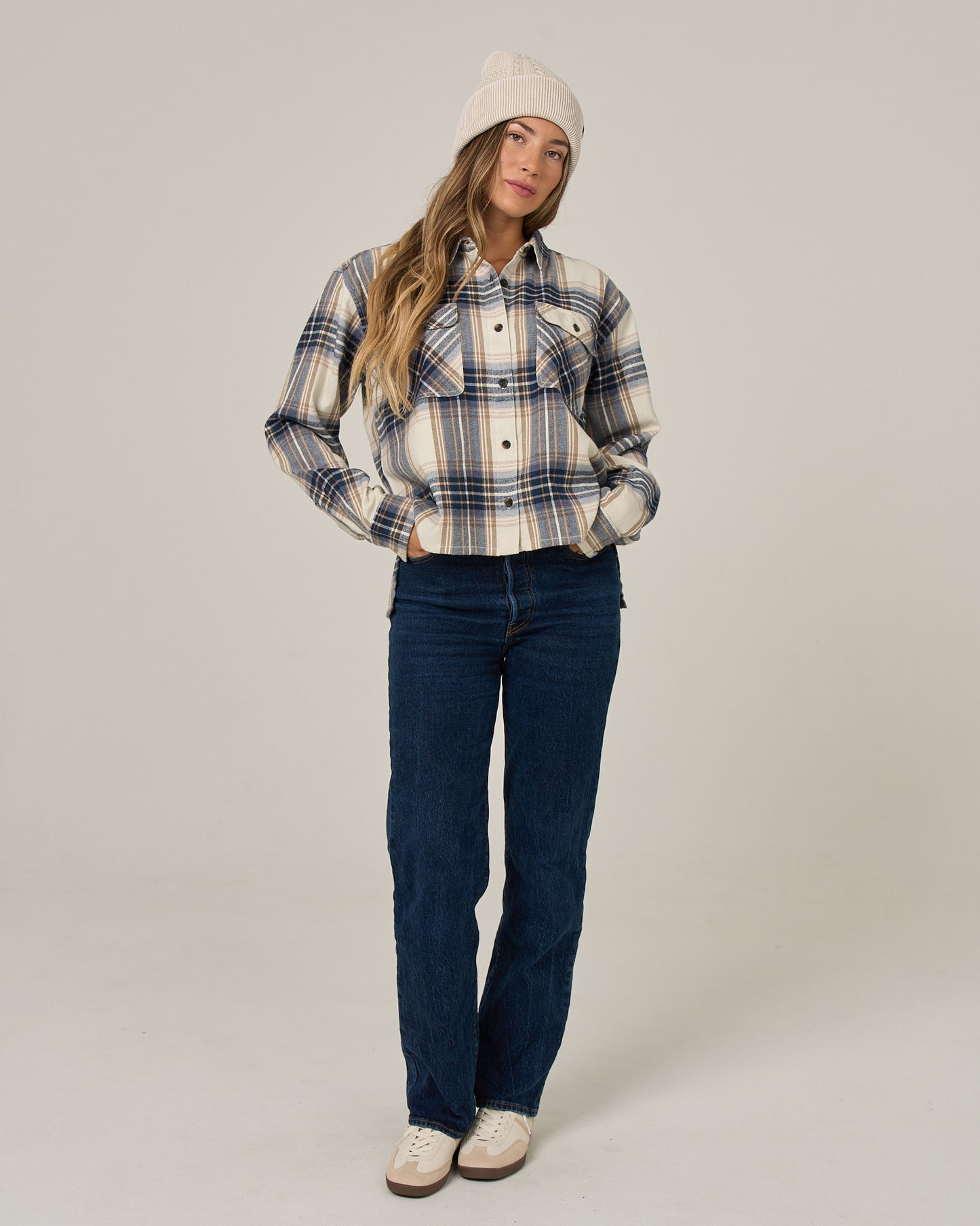 A woman in the Salty Crew Nomad Flannel - Birch, blue jeans, and a beige beanie stands with her hands in her pockets against a plain background.