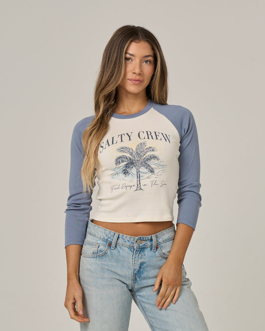 A woman stands facing forward against a neutral background, one hand in her pocket, wearing the Salty Crew Pretty Palms Long Sleeve tee in Flint with a palm tree screen print.