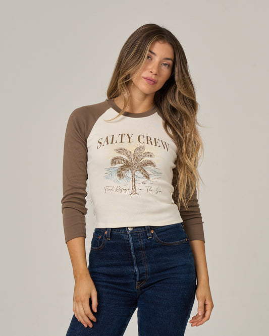 A woman with long hair wears the Salty Crew Pretty Palms Long Sleeve in washed brown, featuring a palm tree graphic, paired with blue jeans, and stands against a plain light background.