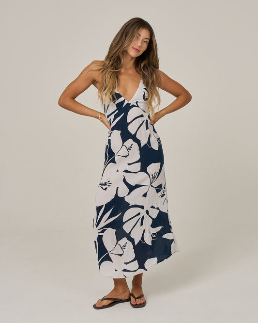 A woman with long wavy hair stands with her hands on her hips against a plain light background, wearing the Salty Crew Graphic Bloom Dress in Faded Mauve—a sleeveless slip dress featuring a deep V-neck, navy and white floral print, cross back straps, and black sandals.