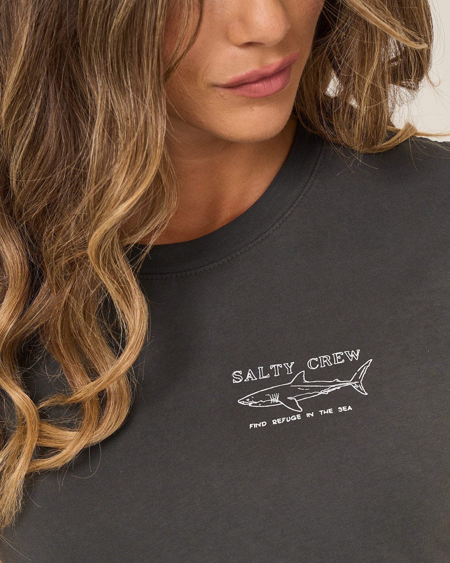 A woman with wavy, light brown hair wears the Salty Crew Shark Anatomy Modern Tee in charcoal, a fitted cotton jersey with cap sleeves featuring a white shark line drawing and "Find Refuge in the Sea.