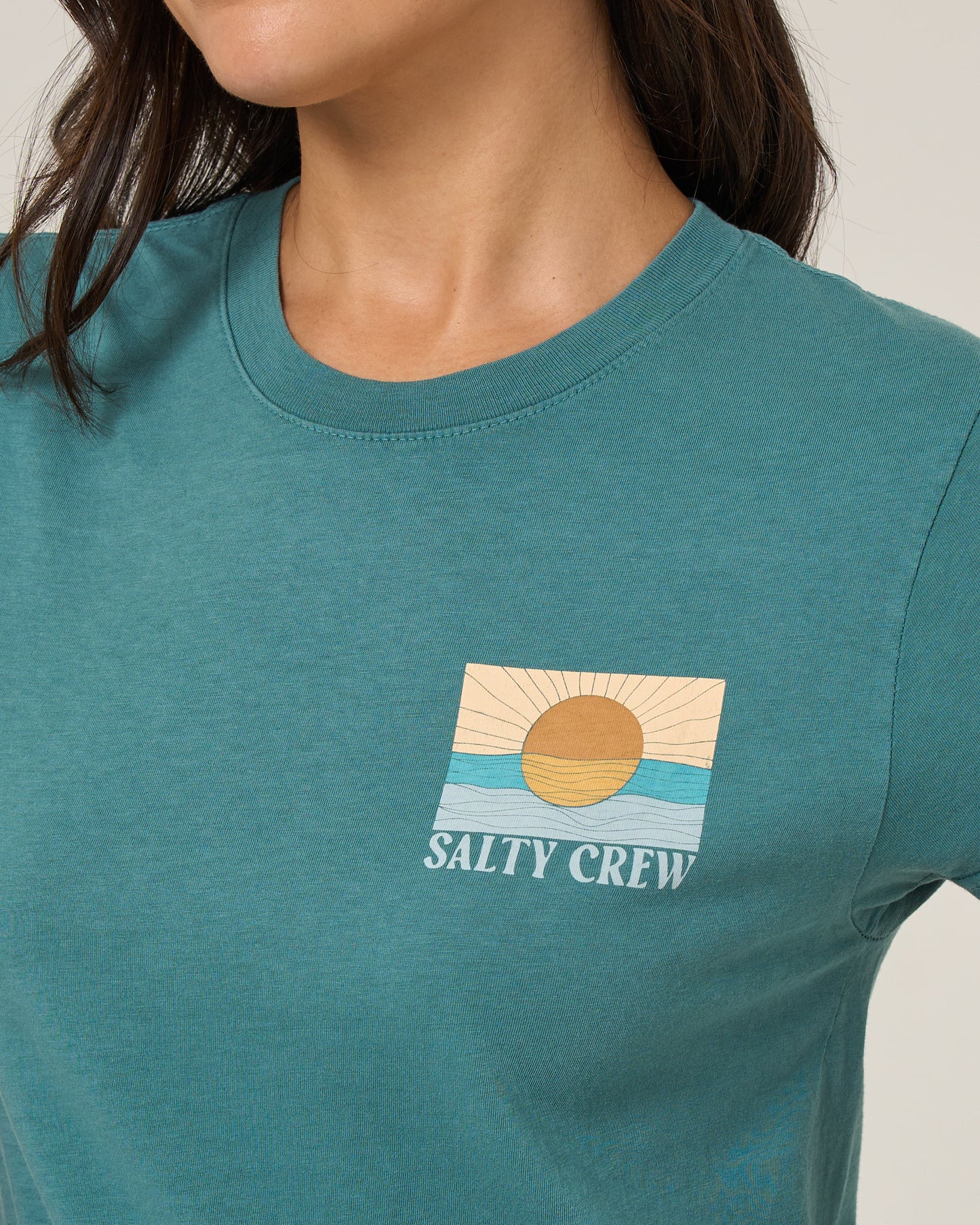 A person wearing the Salty Crew Retro Wave Modern Tee in teal, a cotton jersey shirt with a sun-over-waves screen print and "Salty Crew" below. The image is cropped to show only the upper torso.