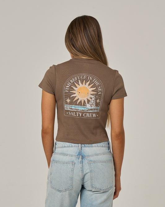 A woman with long, straight hair wears the Salty Crew Celestial Sun Baby Tee in washed brown, featuring a sun and wave graphic with SALTY CREW text on the back. She pairs it with light blue jeans and stands against a plain background.