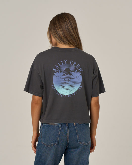 A person is shown from behind against a plain background, wearing the Salty Crew Tropics Skimmer Tee in Charcoal featuring a circular ocean graphic and text, paired with blue jeans.