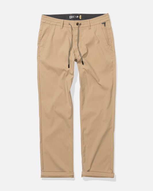 The Transom Tech Pants in khaki are straight-leg pants featuring 4-way stretch, belt loops, a button and zip closure, front and back pockets, and a black drawstring at the waist, shown laid flat on a white background.