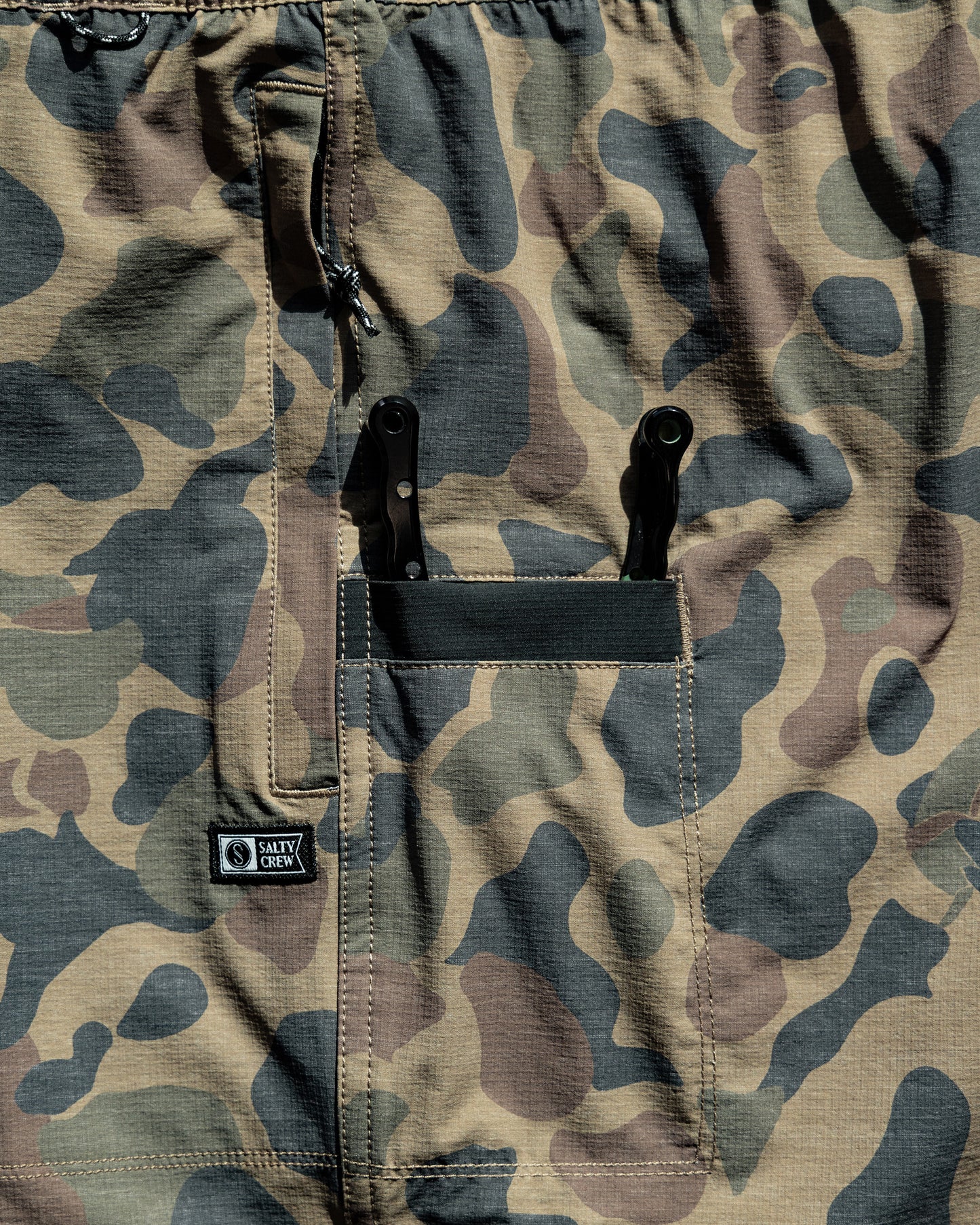 The Salty Crew Crew 18" Ewaist Boatshort in Sand Camo features a durable camouflage-patterned ripstop fabric with a front pocket that holds two multitools and is finished with a stitched black Salty Crew label beside the pocket. Designed for all-day comfort, these shorts offer 4-way stretch and quick-dry properties.