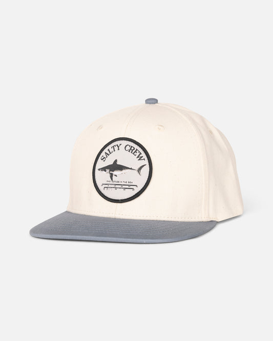 The Salty Crew Bruce Boys 6 Panel in Off White/Slate is a cream cap with a gray brim, featuring a circular woven fish patch and small text on the front. Adjustable snapback closure. Shown against a plain white background.