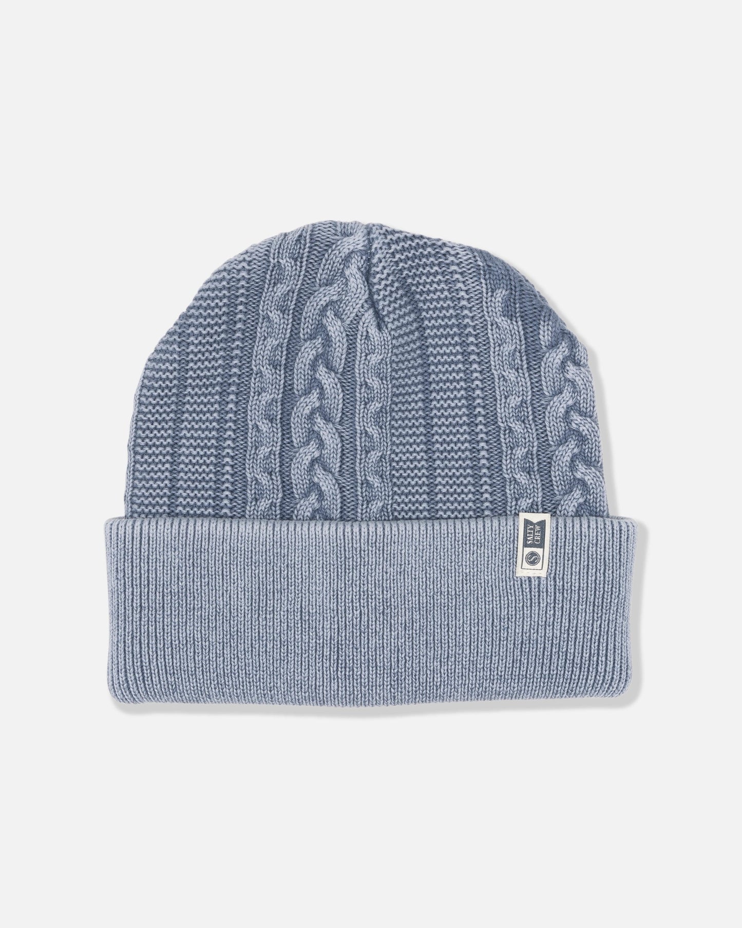The Salty Crew Fisherman Beanie - Flint is a light blue cable knit beanie featuring a folded brim and a vertical cable knit pattern. Crafted in a classic cotton style, it includes a small white rectangular tag with dark text sewn onto the brim. The beanie is shown against a plain white background.