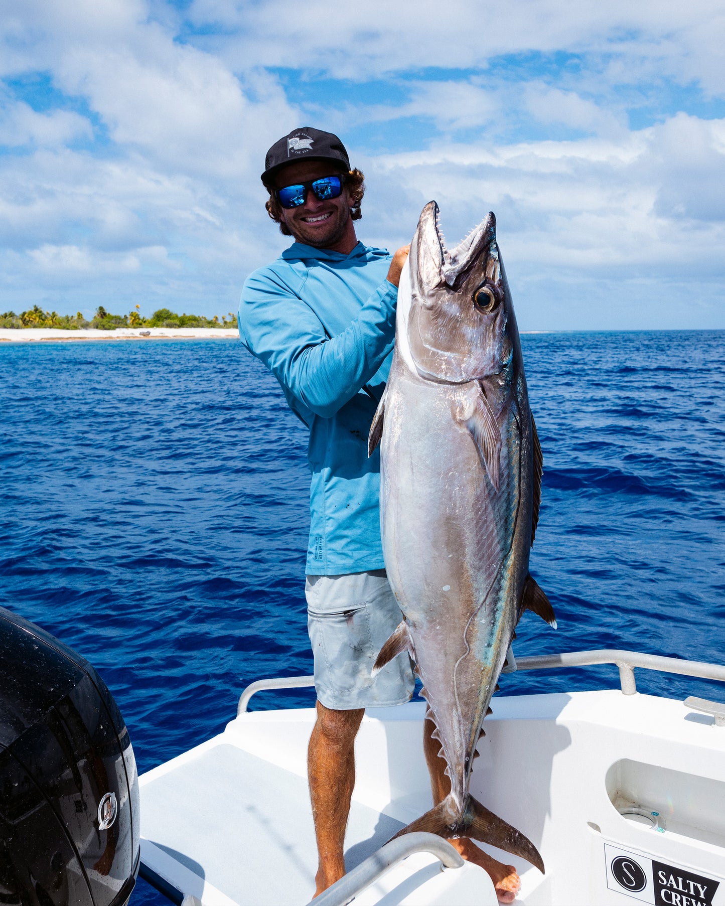 Wearing the Salty Crew Apex Perforated Sunshirt in Captains Blue, a person in sunglasses stands on a boat holding a big tuna, with blue ocean water and a tropical island in the background.