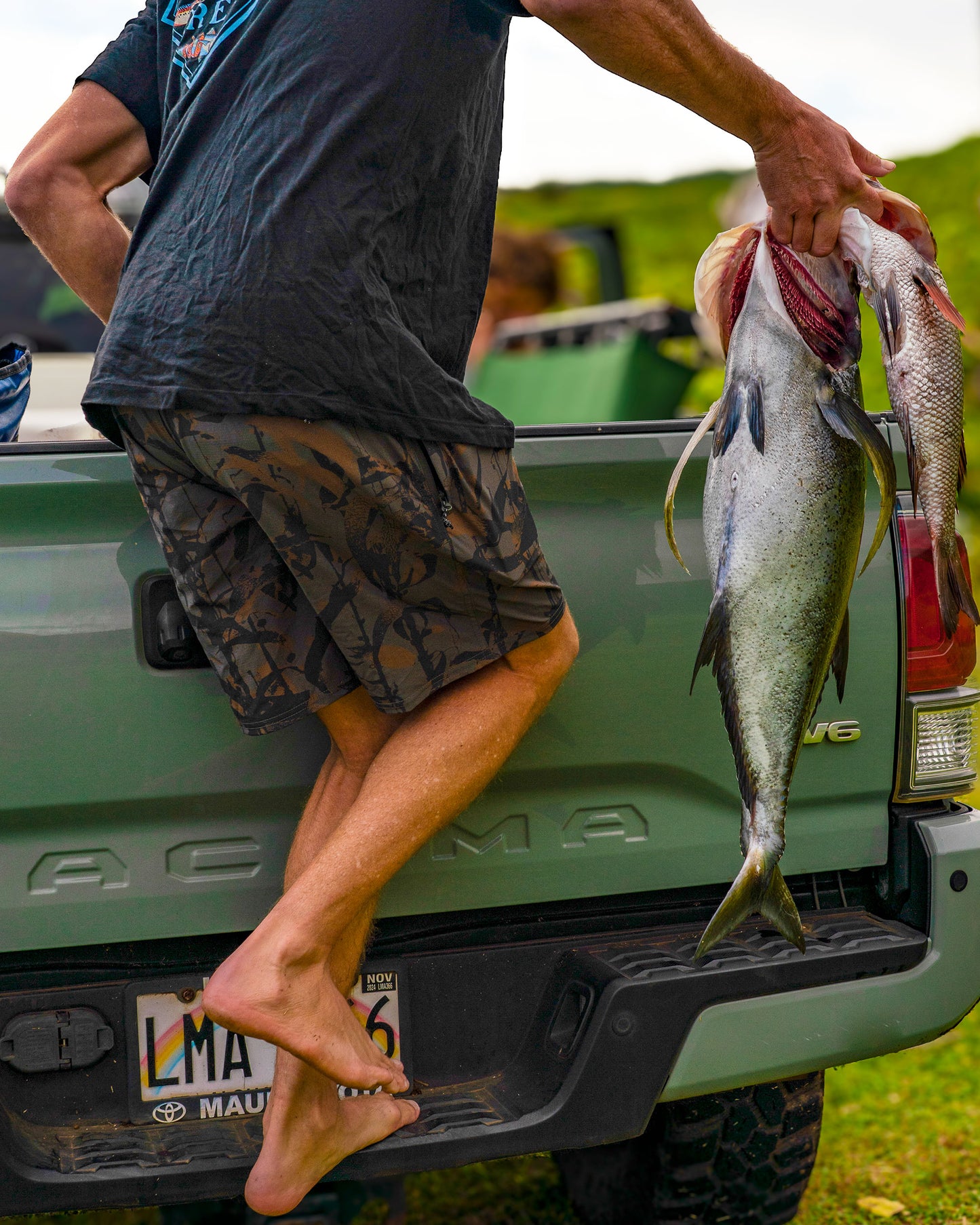 A barefoot person steps onto a truck’s tailgate, holding two large freshly caught fish by the gills. In the bed of the green truck, Salty Crew Crew 18" Ewaist Boatshorts in Dark Kelp Camo—made from quick-dry ripstop fabric with 4-way stretch—are tossed alongside gear bags, ready for adventure.