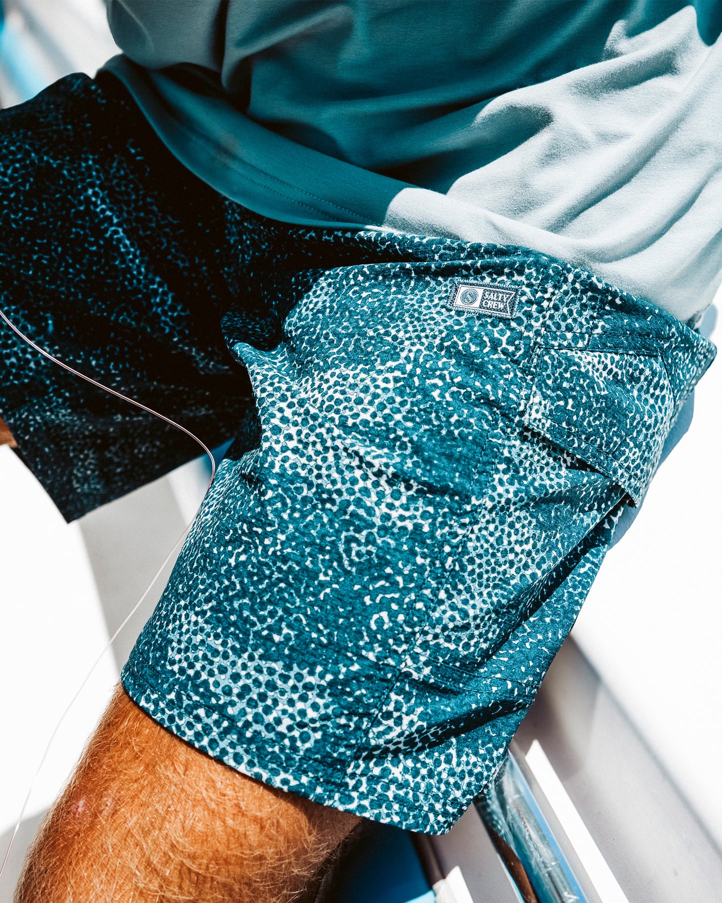 A person sits on a white bench in bright sunlight, wearing Salty Crew’s Flagship 20" Boatshort - Oceanic, featuring a blue and white pattern and quick-dry fabric, paired with a light blue shirt.