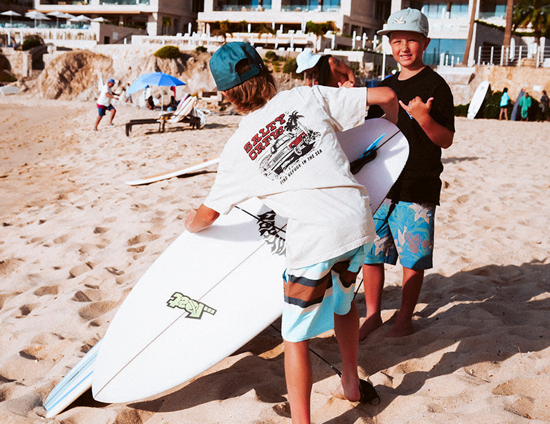 Two boys wearing shorts and t-shirts stand on a sandy beach. One holds a surfboard while the other gives a thumbs up. People and beach umbrellas are visible in the background near beachfront buildings.