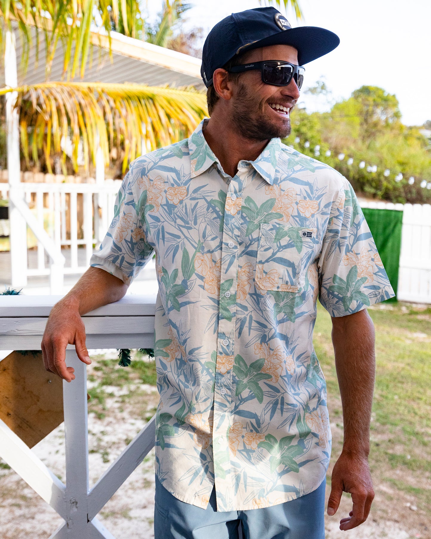 A man in the Salty Crew Crew Button Up - Off White, sunglasses, and a black cap stands outdoors, smiling and leaning on a white wooden railing against a tropical backdrop.