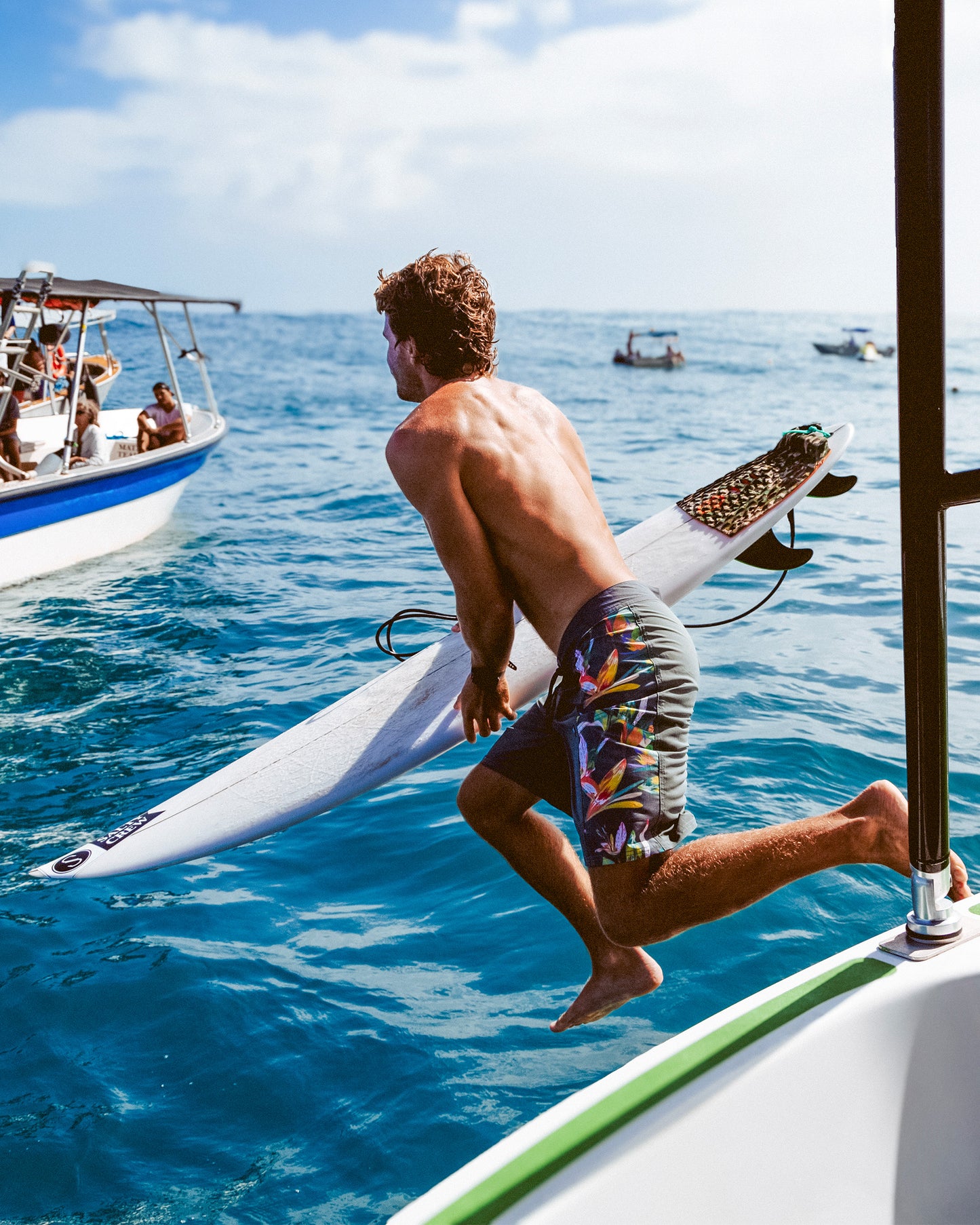 Wearing Salty Crew's Flagship 20" Boardshort in Washed Green, a man leaps off a boat into the ocean with his surfboard. Other boats and people are seen under a bright, partly cloudy sky in the background.