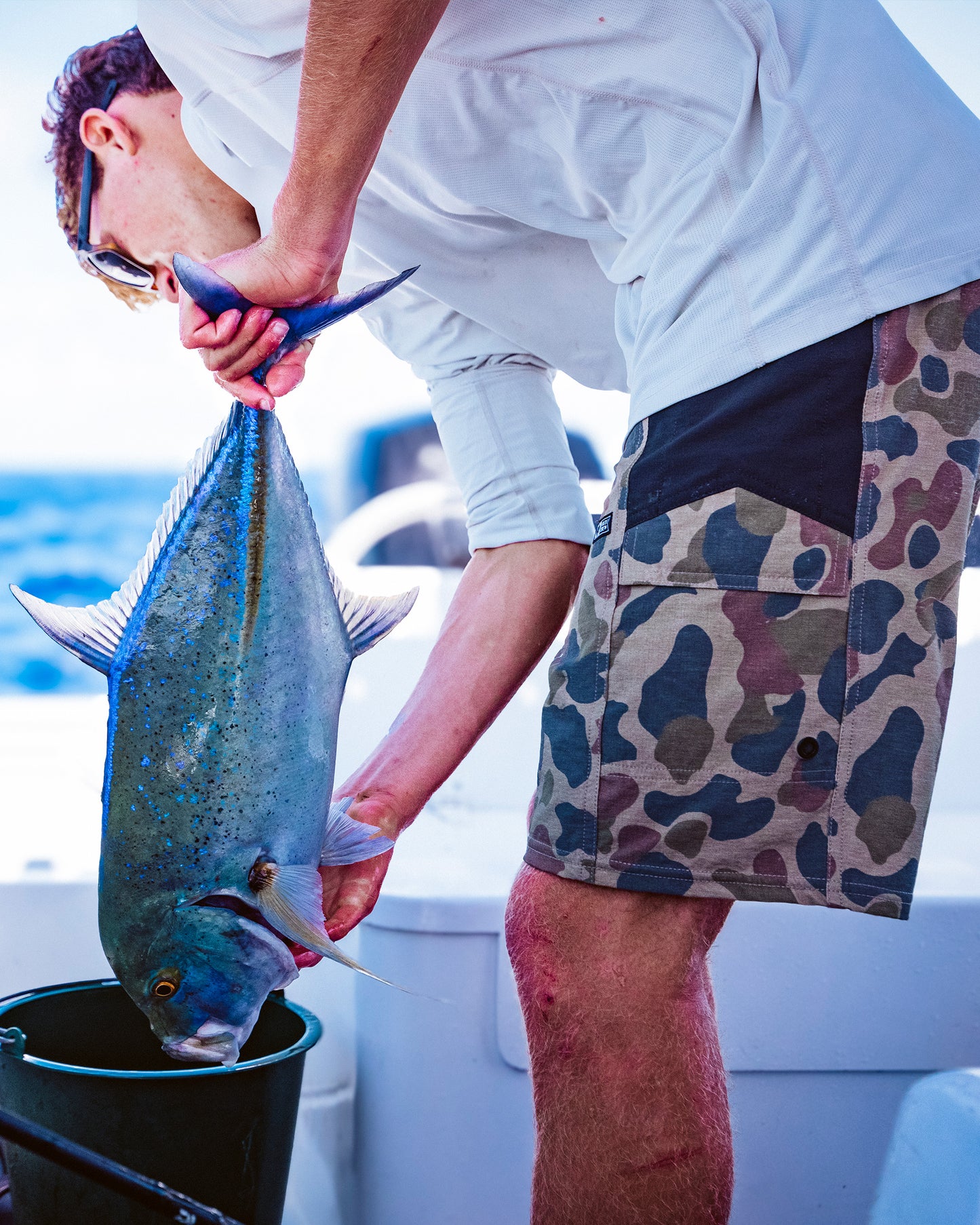 Wearing Salty Crew’s Flagship 20" Boatshort in Sand Camo with quick-dry ripstop fabric, an angler in a white shirt holds a large fish by the tail over a bucket on a boat during a sunny fishing trip.