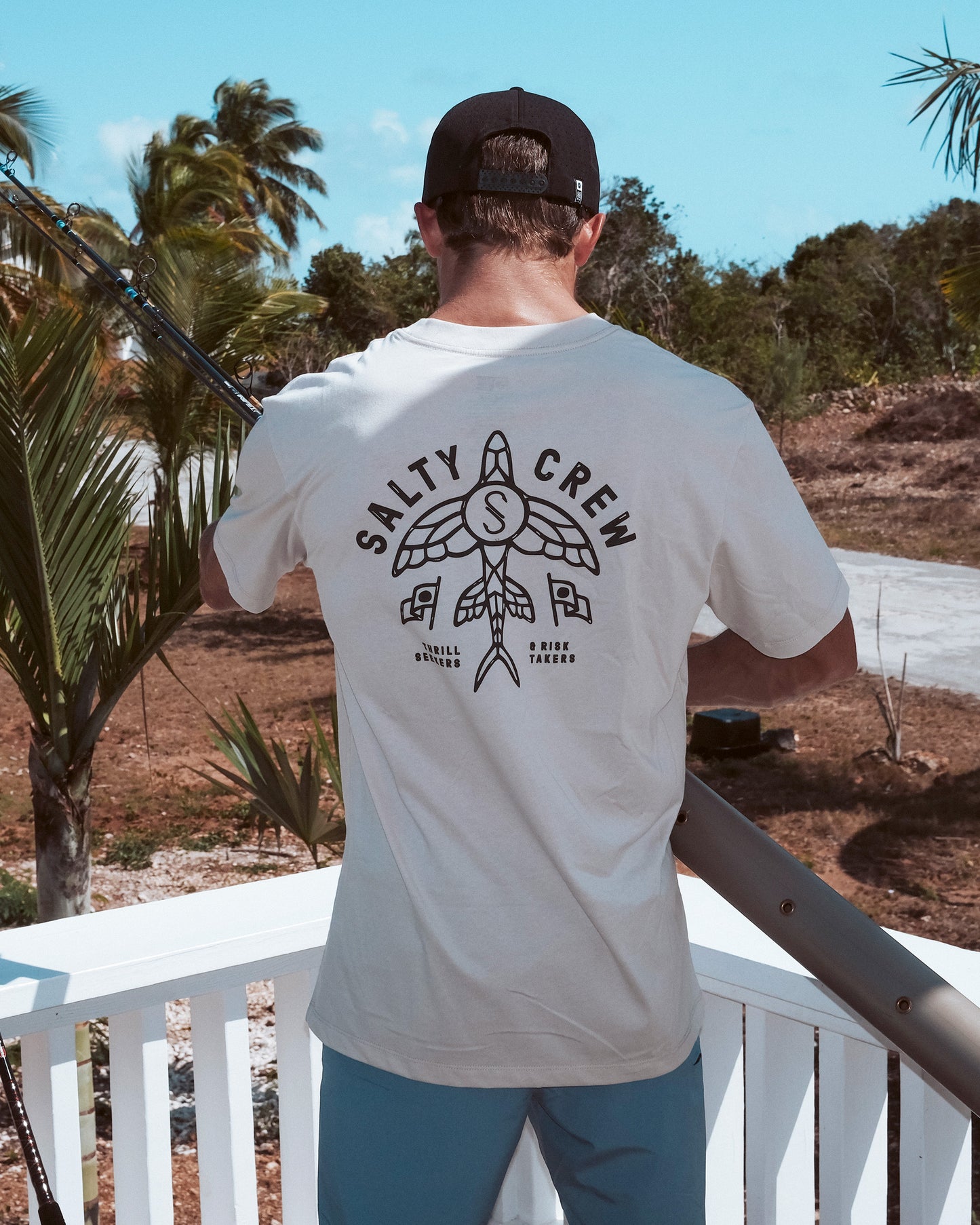 A person stands on a porch facing away, holding a fishing rod and wearing the Salty Crew Flyer Tee - Bone. The white T-shirt features a screenprinted graphic on the back, with palm trees and blue sky in the background.