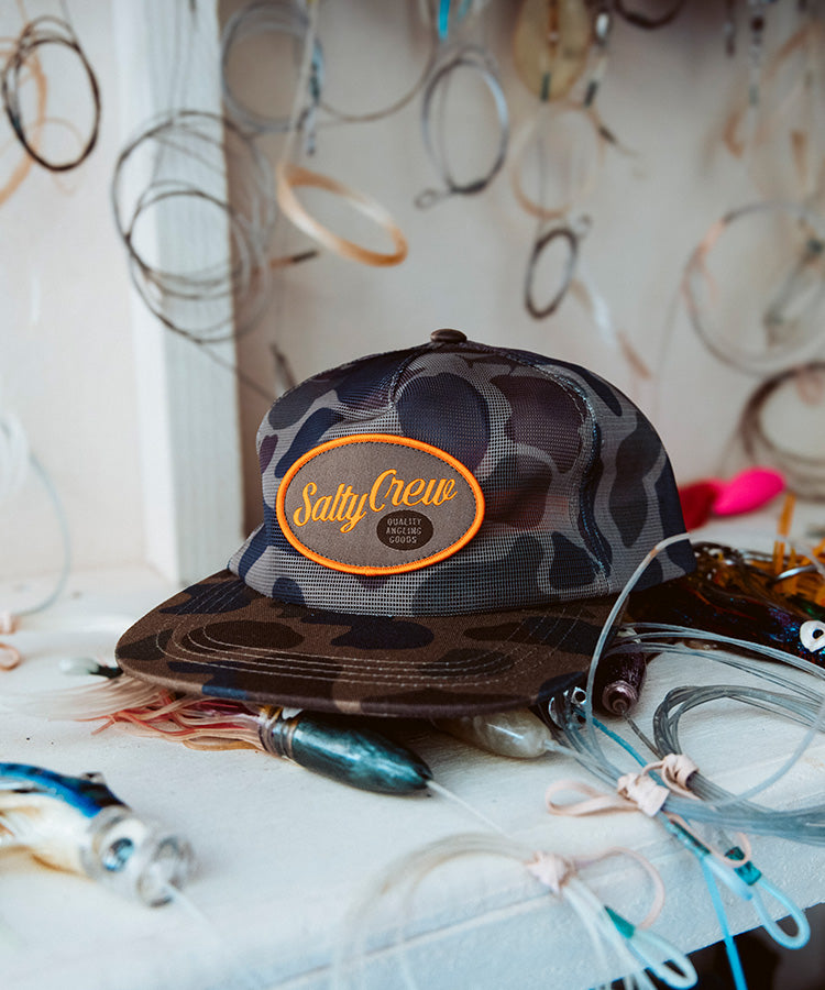 The Salty Crew Netting Mesh Trucker - Sand Camo sits on a table with fishing lures, hooks, tangled lines, and other gear hanging in the background.