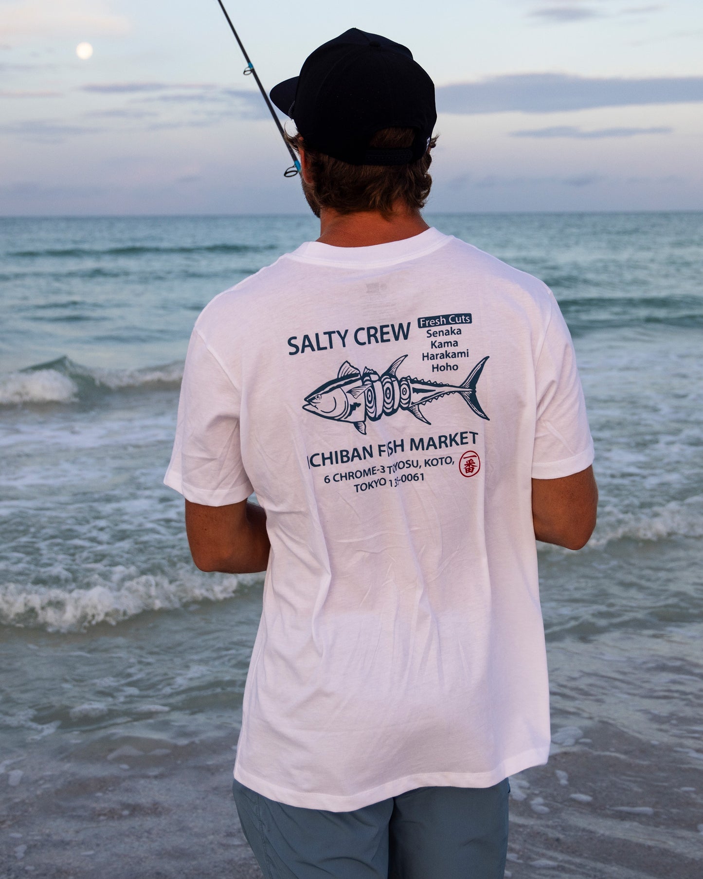 A person in a white Salty Crew Ichiban Tee with a fish graphic stands on a sandy beach, facing the ocean and holding a fishing rod under a cloudy sky.