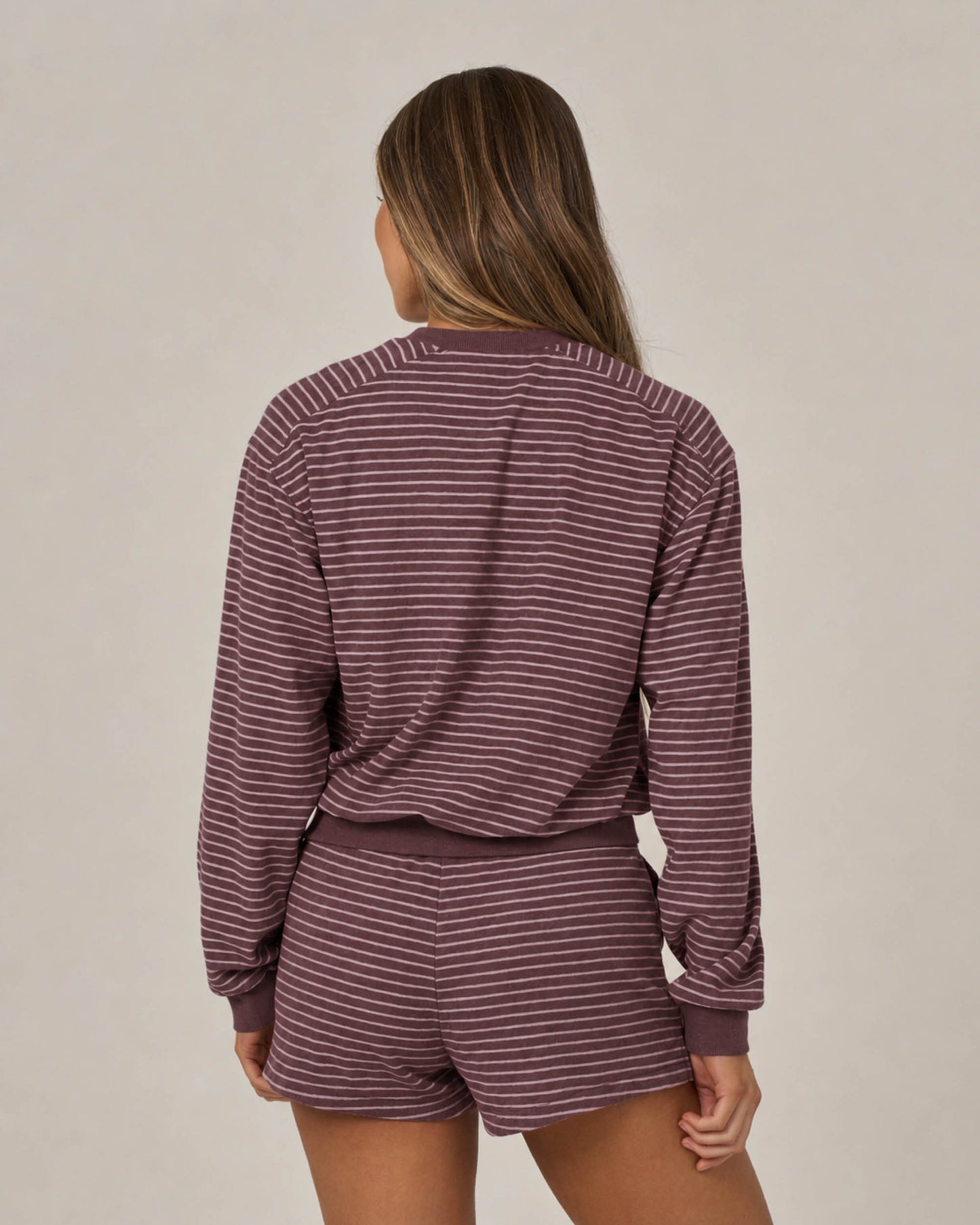 A person with long hair is standing with their back to the camera, wearing the Salty Crew Leeward Crew in maroon—a matching maroon and white stripe crew neck long-sleeve top and shorts set featuring a branded patch—against a plain background.
