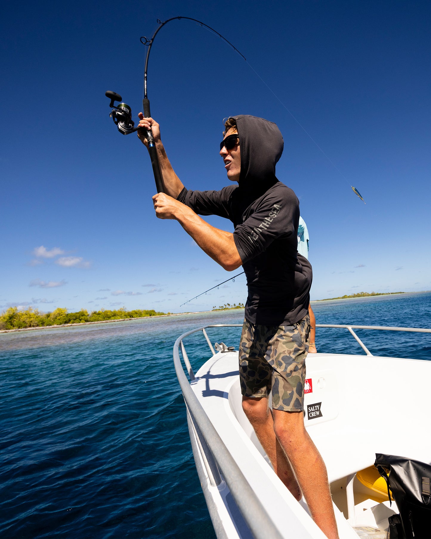 Wearing the Salty Crew Mariner Lite UV Hood in Charcoal, offering UPF 50+ protection, a person stands on a white boat holding a fishing rod over blue ocean water near a tropical shore under clear skies.