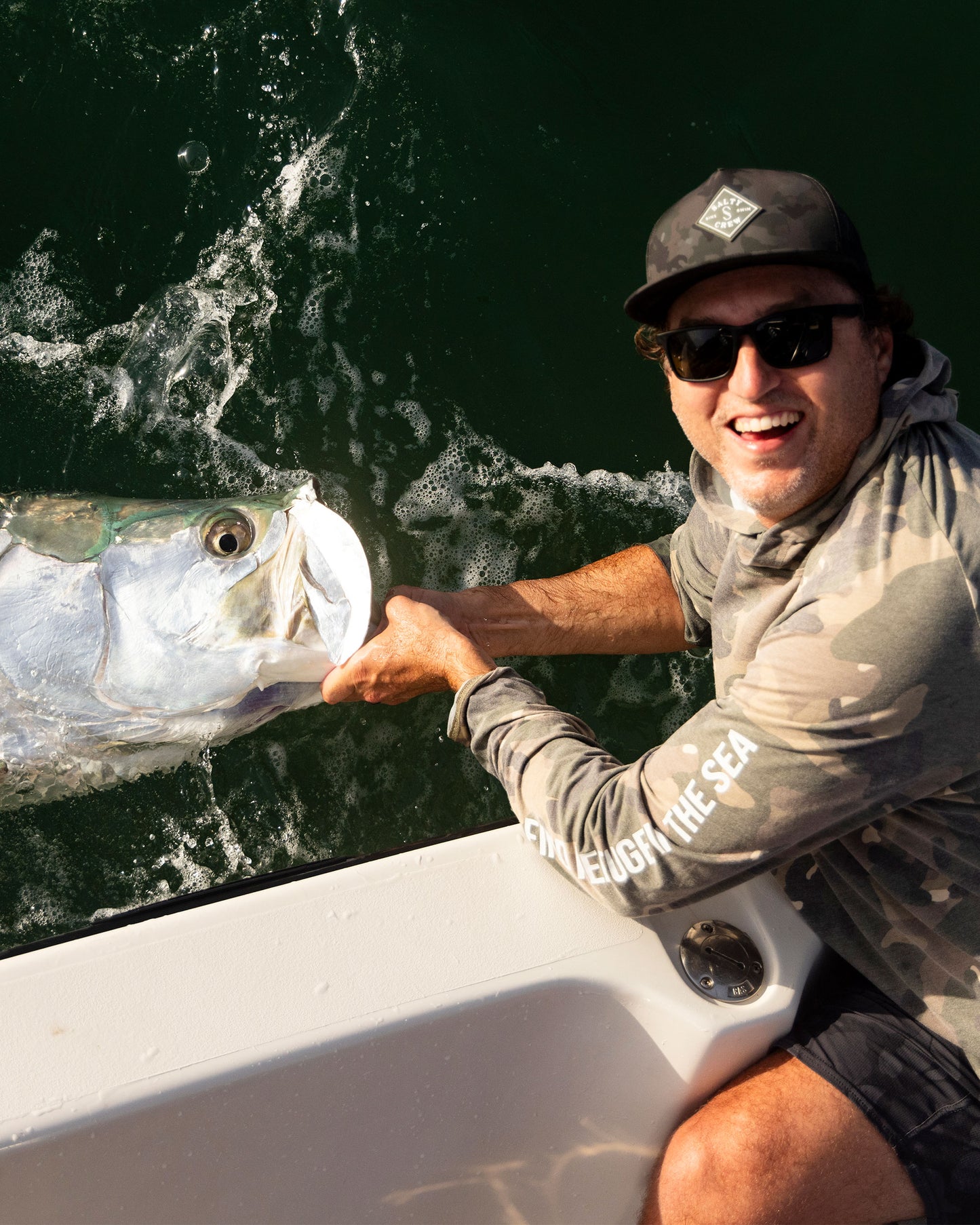 A man in sunglasses, a cap, and a Salty Crew Mariner Lite UV Hoodie - Green Camo with UPF 50+ grins as he holds a large fish by the mouth over a boat side above green water.