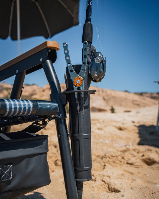 A Parkit x Salty Crew Voyager Accessory Mount by PARKIT Co. secures a fishing rod to a beach chair, with a striped towel draped over the chair and sand plus blue sky in the background.