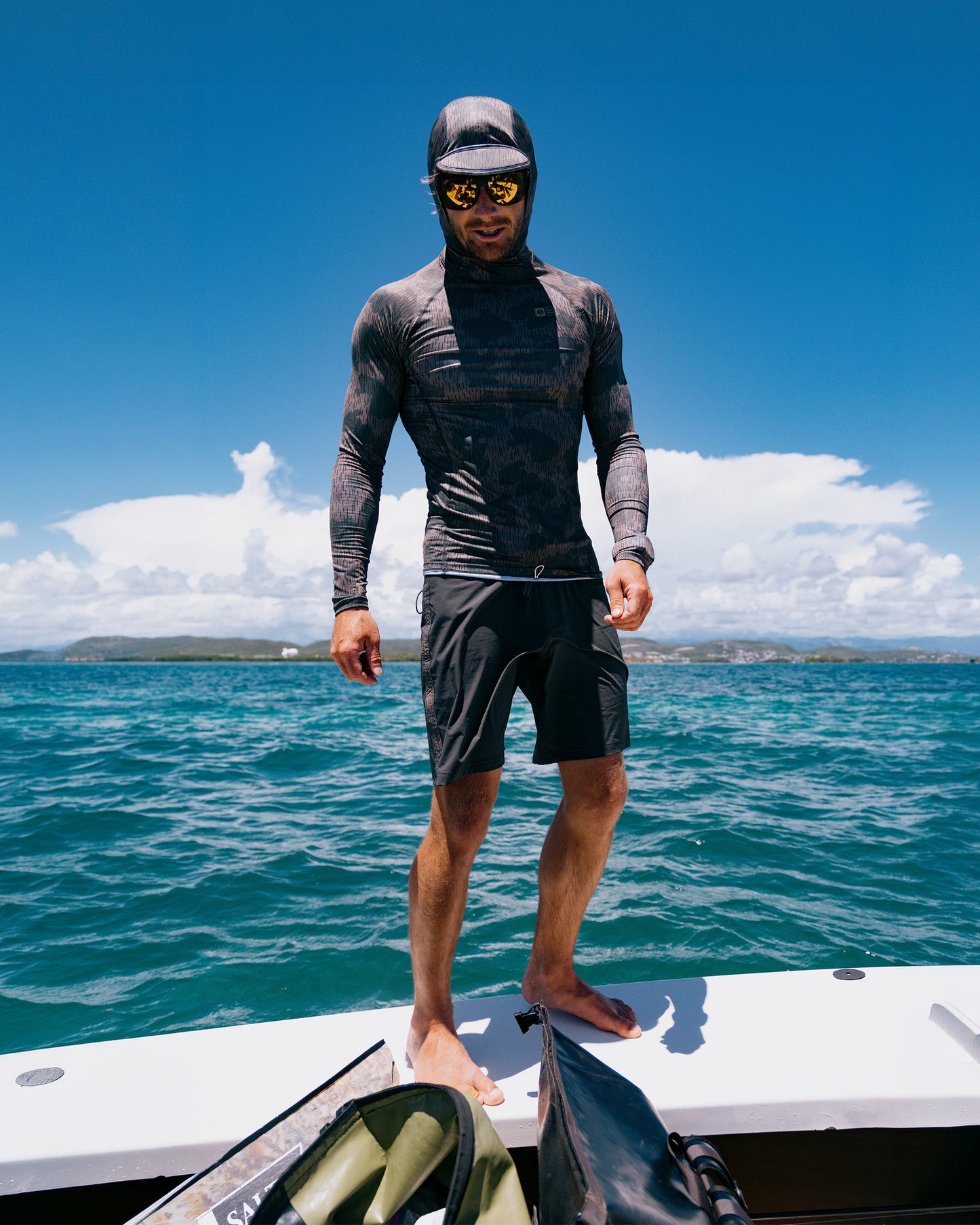 A man in a Salty Crew Apex Hooded Rashguard - Recon Camo with UPF 50+, shorts, and sunglasses stands barefoot on a boat under sunny skies, blue ocean and distant land visible in the background.