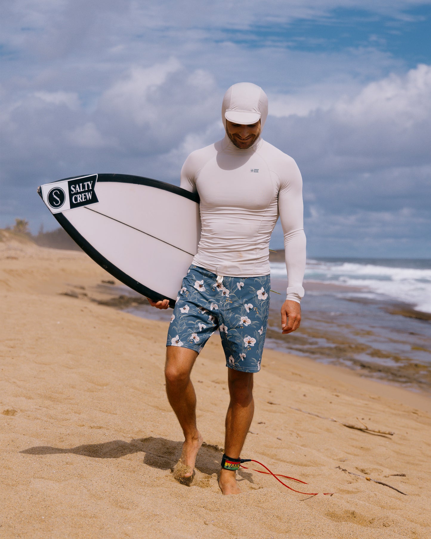 Wearing a Salty Crew Apex Hooded Rashguard in light grey and patterned swim trunks, a person walks barefoot on the sand holding a Salty Crew surfboard as waves roll in beneath a partly cloudy sky.