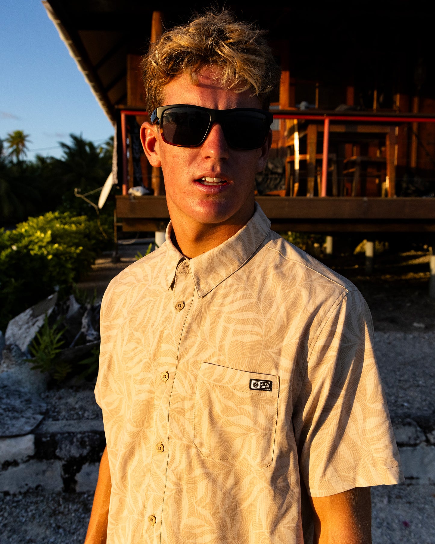 A young man with tousled blond hair and dark sunglasses stands outdoors at sunset wearing the Salty Crew Breezer Perforated Button Up in Khaki, a lightweight, breathable short-sleeve shirt. A wooden building and greenery are in the background.