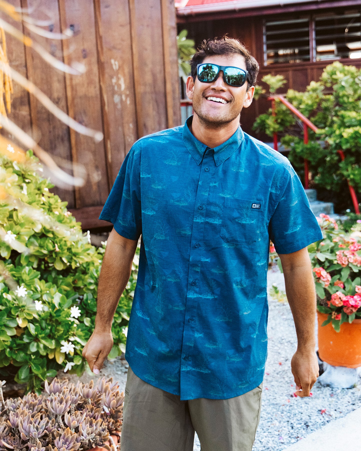 A man wearing the Salty Crew Breezer Perforated Button Up in navy stands outside, smiling amid greenery and flowers with a wooden building behind him. The lightweight, breathable shirt is perfect for warm weather.