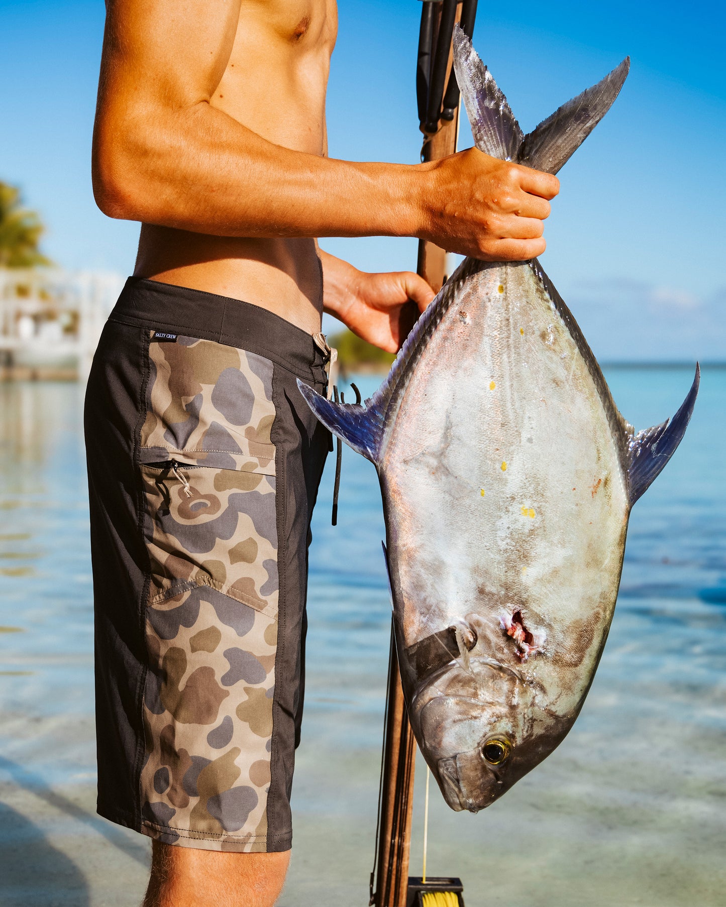 Wearing Salty Crew's Flagship 20" Boardshort in Sand Camo, a person stands in shallow water holding a large fish by the tail, with a tropical beach and clear blue sky in the background.