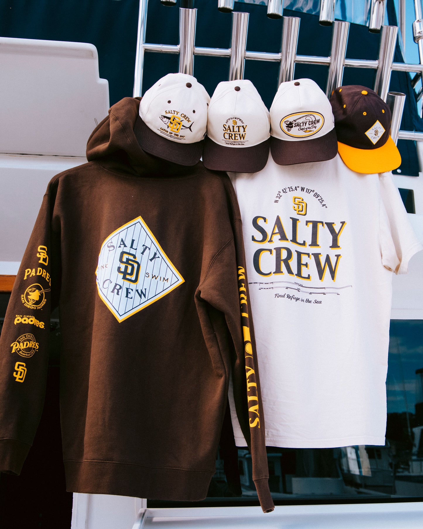 Brown hoodie, cream t-shirt, and four hats—including the Padres Tippet Hat in brown by Salty Crew—hang on a white railing, displayed on a boat. Both shirts and hats feature yellow, brown, and white color schemes.