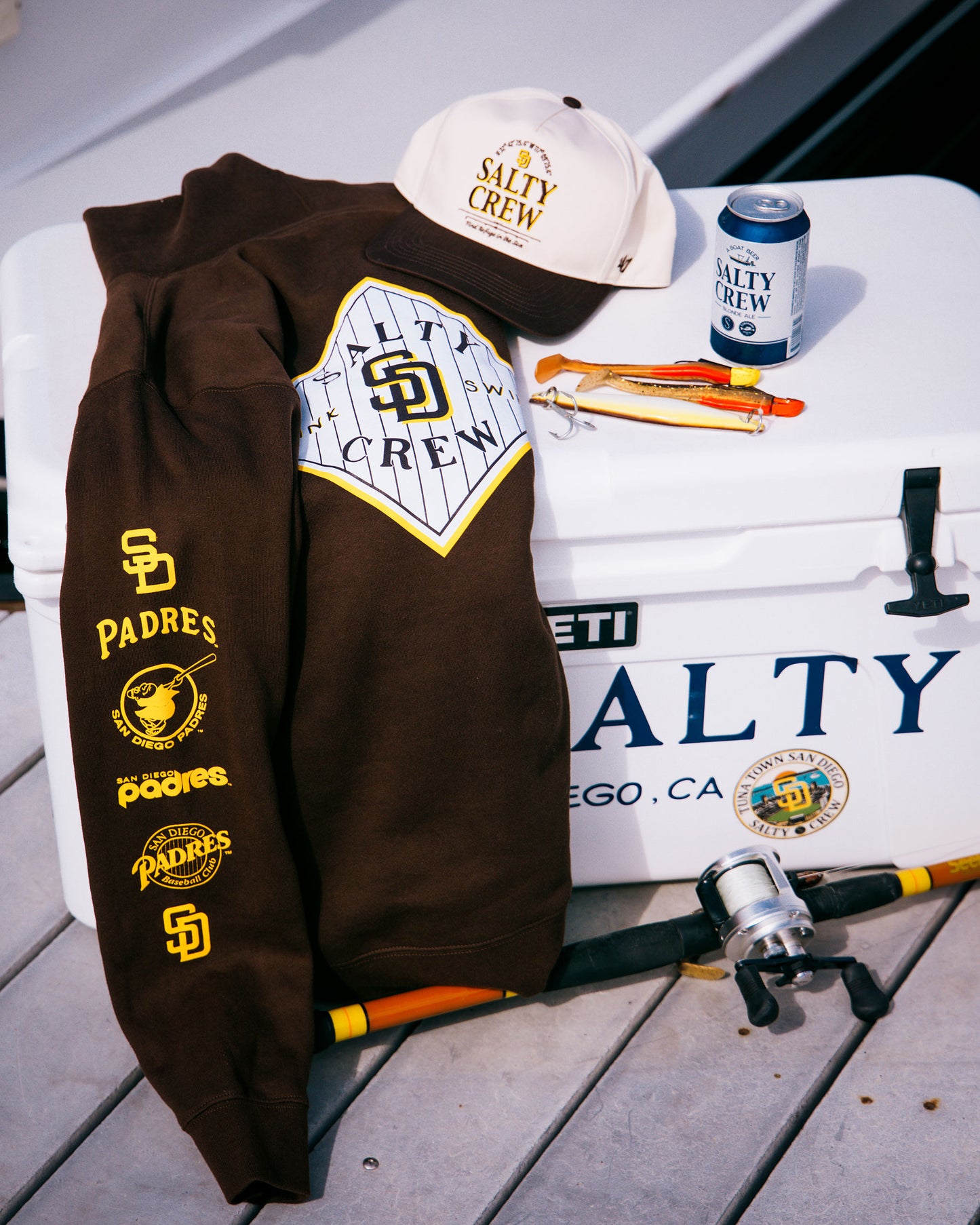 A limited edition brown Padres hoodie, a Salty Crew Padres Tippet Hat in Bone, a Salty Crew cap, can, fishing pliers, and a fishing pole rest on a white YETI cooler labeled Salty Crew on a dock.