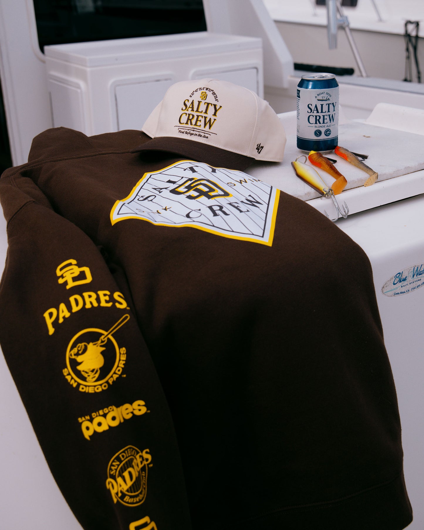 A white Padres Catch of the Day Tee from Salty Crew, a can, and a plate with cheese and crackers rest on a white surface, possibly on a boat.