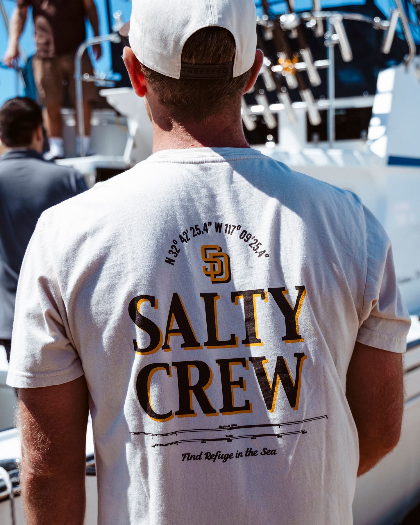 A man wearing the Salty Crew Padres Label Tee in Bone and a cap stands on a boat dock, with other people and a boat visible in the background. The t-shirt, inspired by the San Diego Padres, features coordinates and “Find Refuge in the Sea” printed on it.