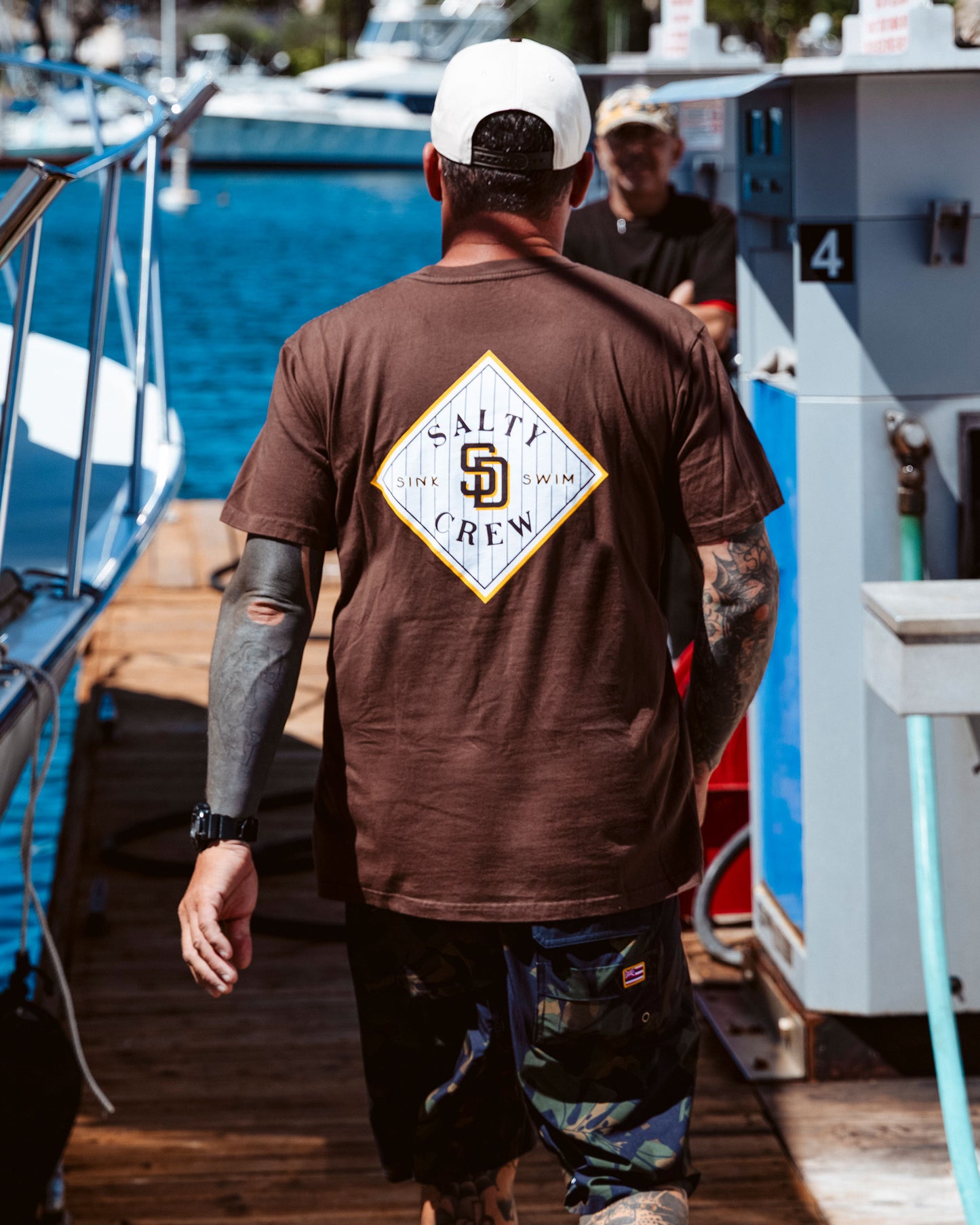 A man wearing a limited edition Salty Crew Padres Tippet Tee in brown and camouflage shorts stands on a dock near a boat and gas pump, with water and more boats in the background.