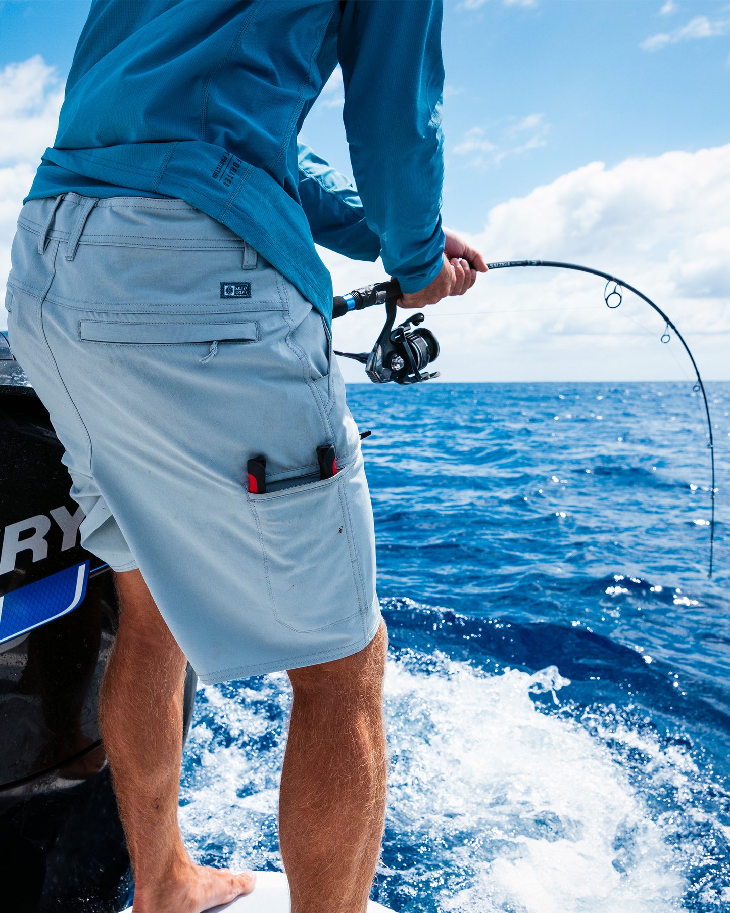 A person wearing Salty Crew’s Transom 19" Tech Short in Quarry and a blue shirt stands barefoot on a boat, fishing with a rod over the deep blue ocean beneath a partly cloudy sky.