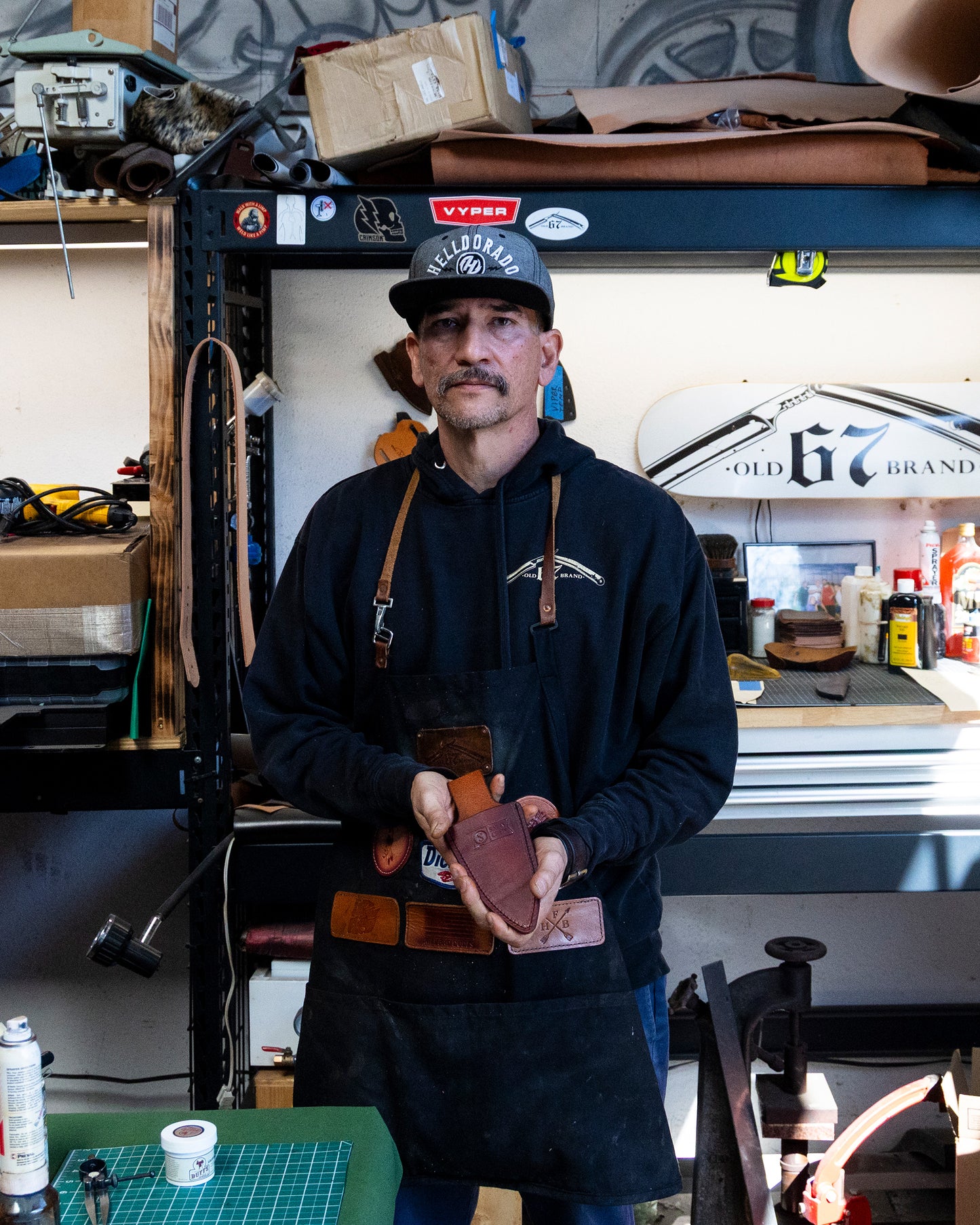 A man in a black cap, hoodie, and apron stands in a workshop holding the Salty Crew Old 67 x Salty Crew Plier Holster. Shelves with tools, boxes, bottles, and a skateboard labeled Old 67 Brand are visible behind him.