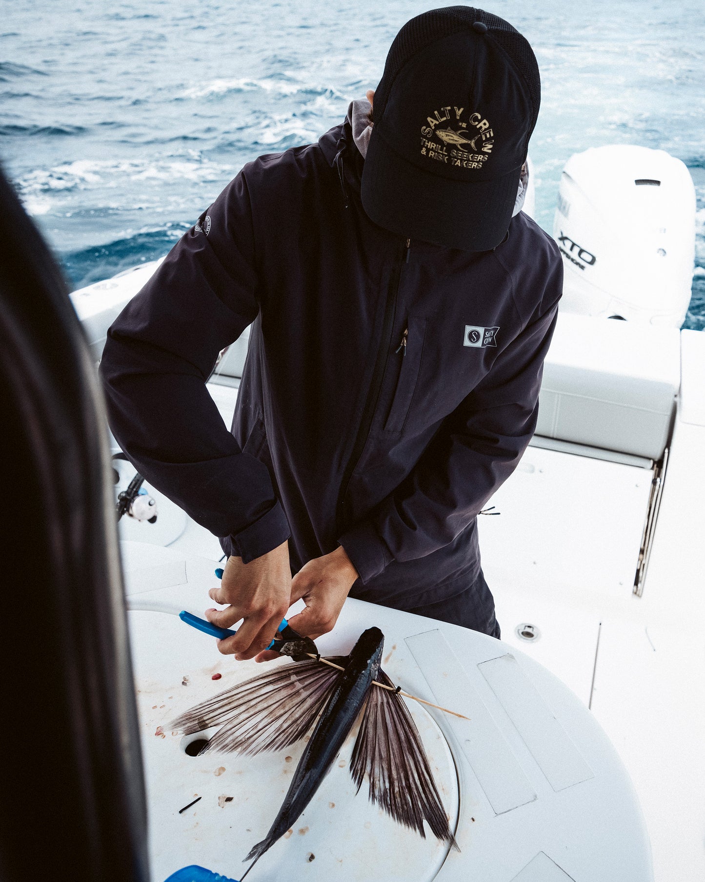 Wearing the Salty Crew Stamped Trucker - Black, a person fillets a long fish with a sharp knife on a round cutting board aboard a boat, the ocean visible in the background.