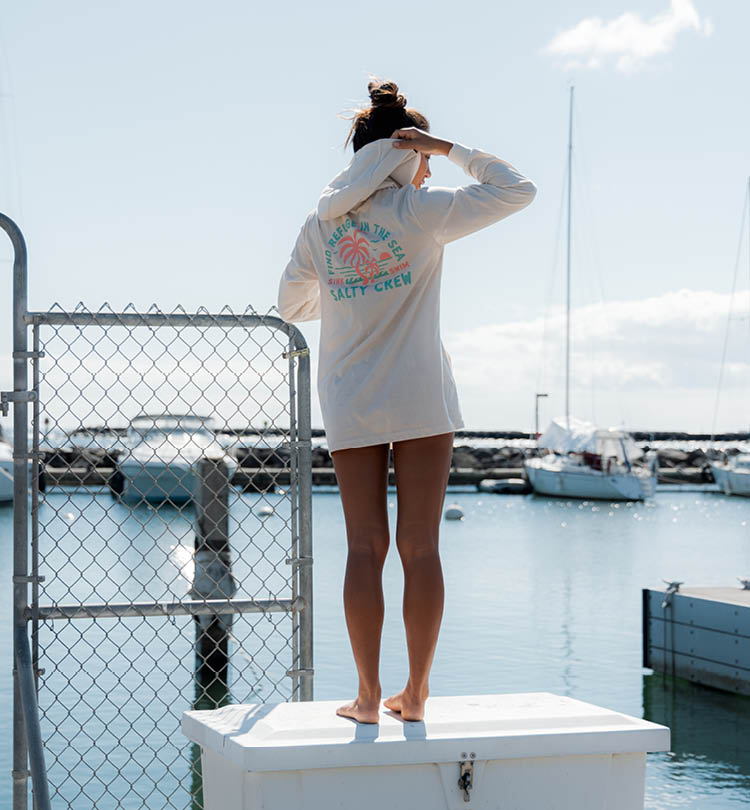 A person stands barefoot on a white box by a marina, facing the water. They wear a long-sleeve shirt with a colorful back print and hold their hands behind their head. Boats and a chain-link fence are in the background.