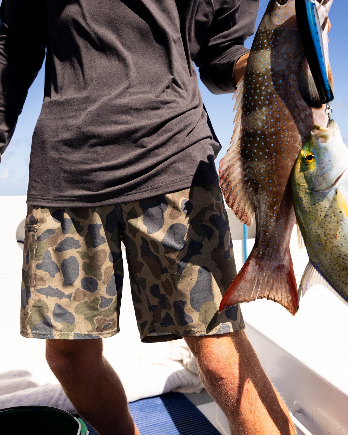 A person in a black shirt and Salty Crew Transom 19" Tech Short - Sand Camo stands on a boat, holding up two freshly caught fish with one hand on a sunny day.
