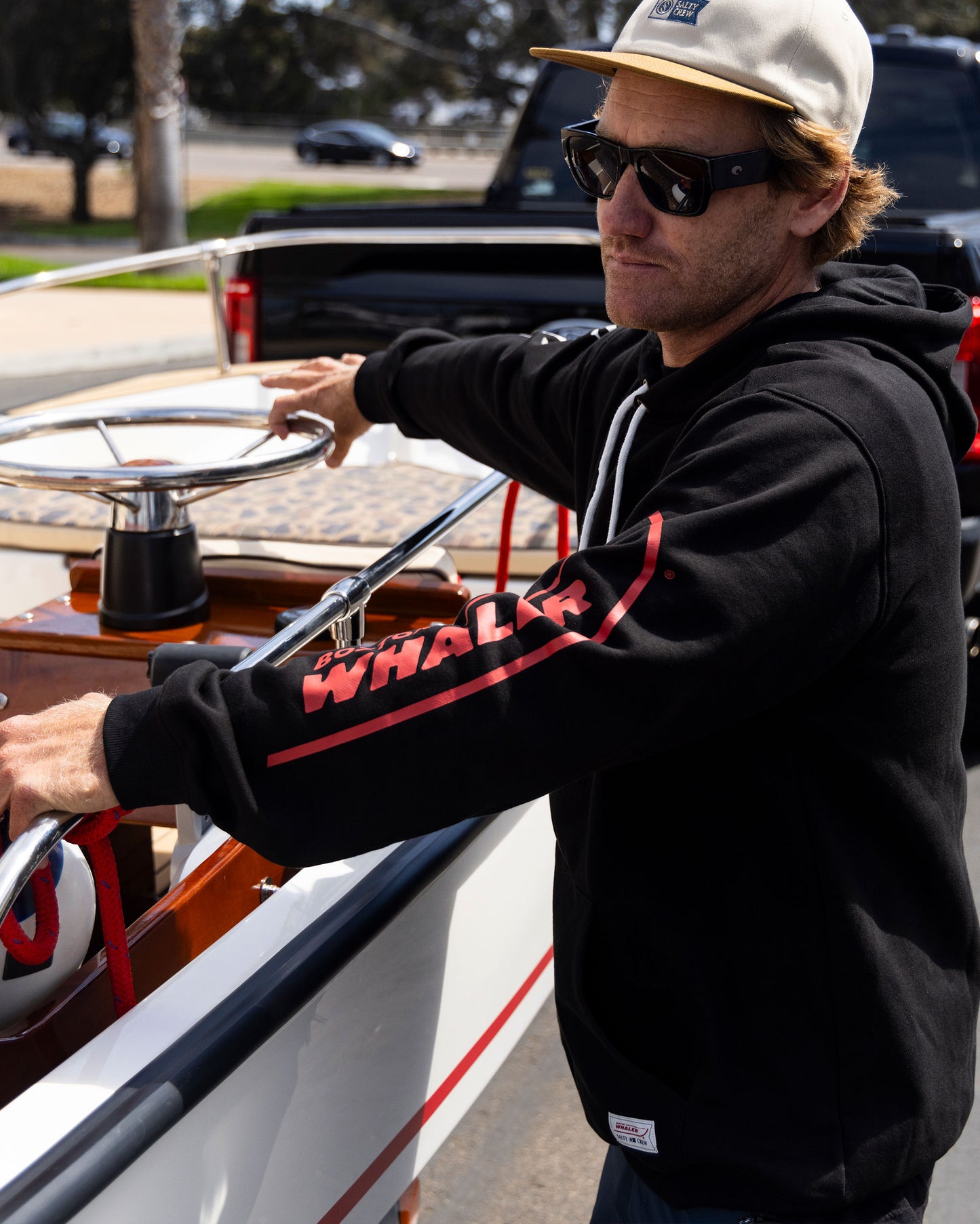 A man in Salty Crew’s Whaler Icon Fleece - Black, sunglasses, and a tan cap stands by a boat, gripping the steering wheel on a sunny day. The black cotton-poly hoodie features screenprinted "WHALER" on the sleeves.