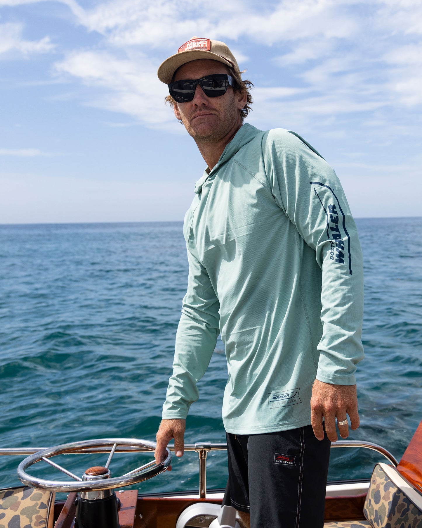 A man in sunglasses and a cap stands at the wheel of a boat, wearing the Salty Crew Whaler Apex Sunshirt - Mackerel, a light blue UPF 50+ long-sleeve, with blue ocean water and partly cloudy sky in the background.