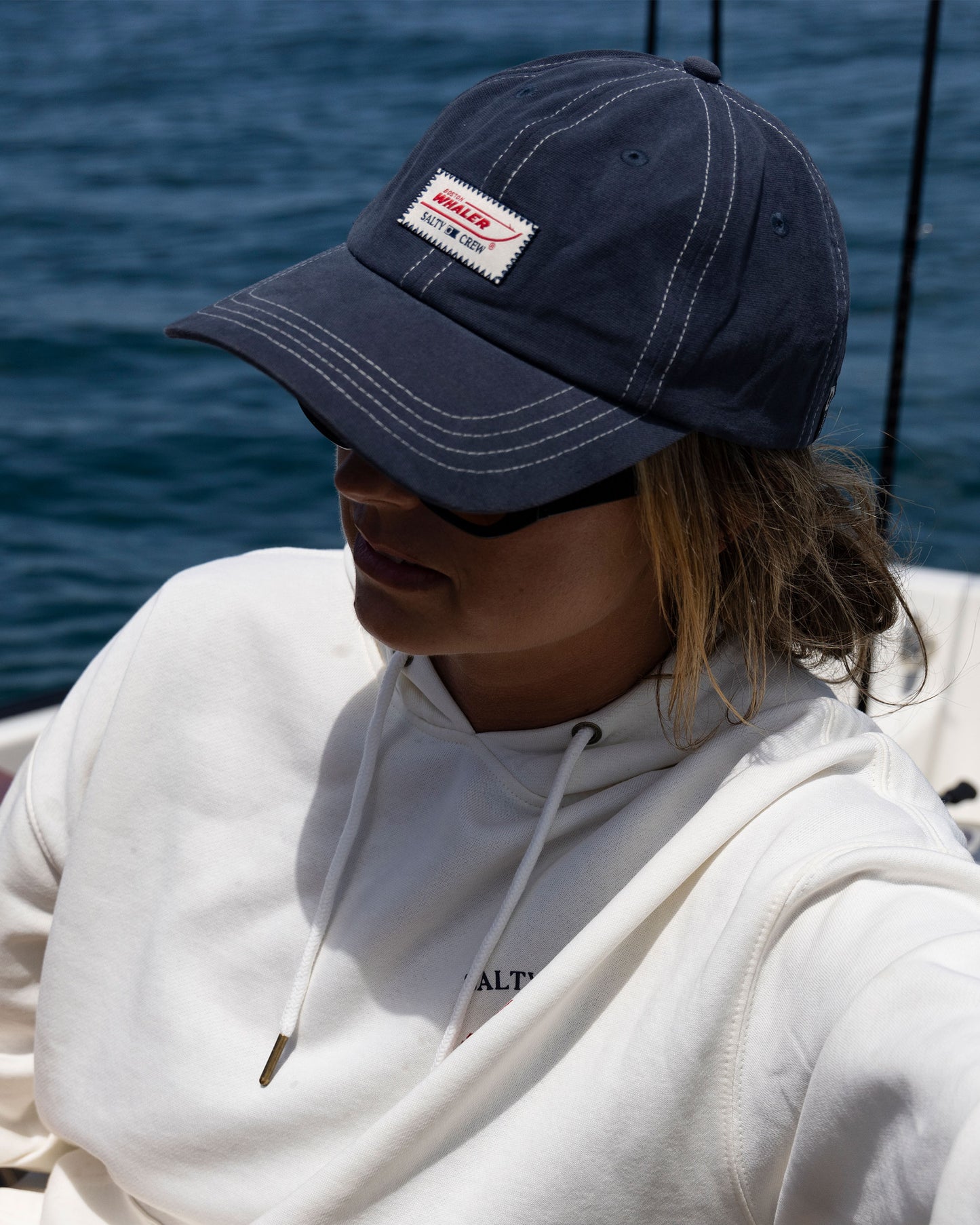 A person wearing a Salty Crew Whaler Dad Hat in Midnight Navy, sunglasses, and a white hoodie sits on a boat with water behind them. The hat features a patch with text on the front.