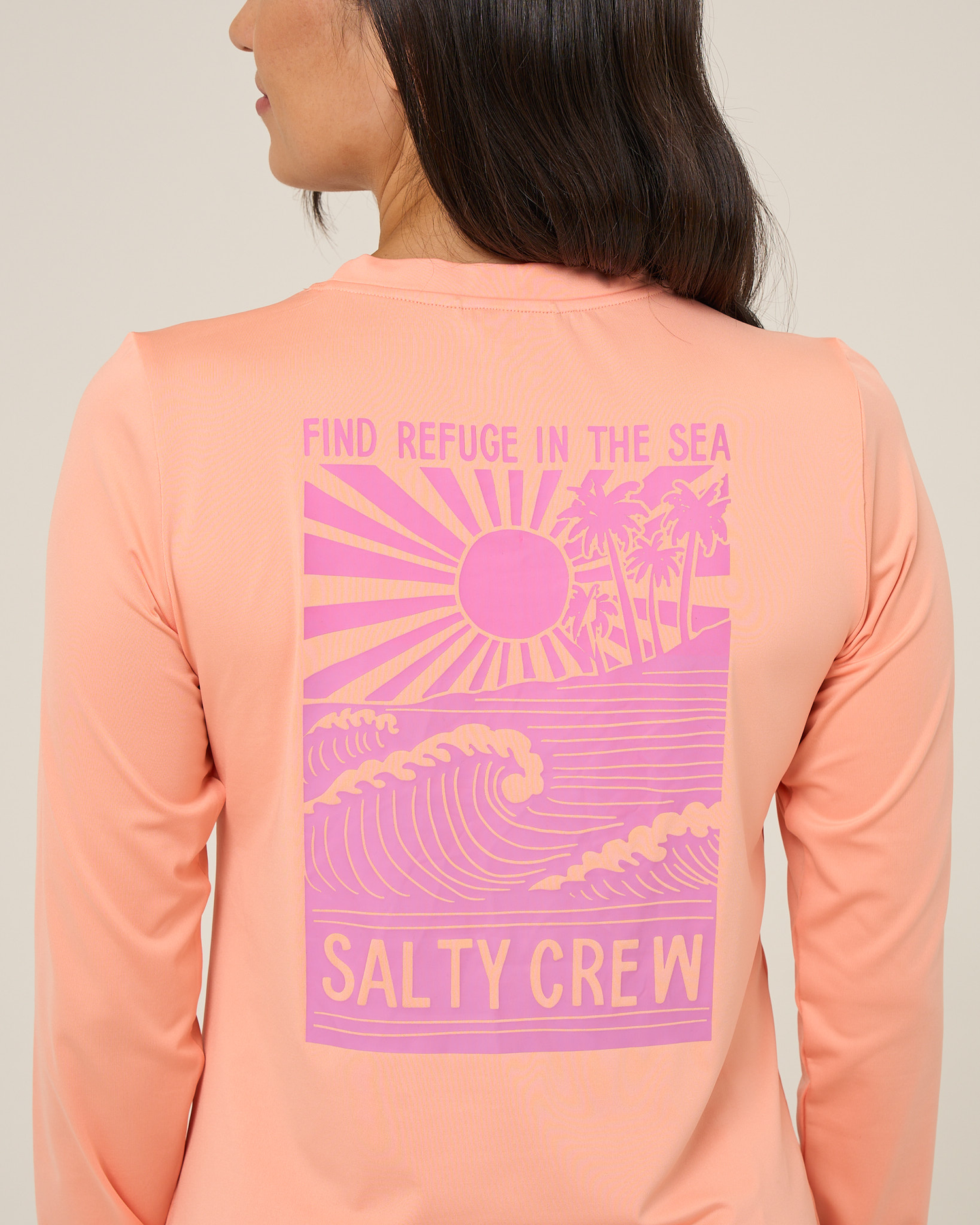 A person wearing the Salty Crew Rise And Smile Sunshirt in Papaya, featuring UPF 50+ protection and a pink graphic of waves, sun, and palm trees with the words "FIND REFUGE IN THE SEA SALTY CREW." This moisture-wicking shirt is shown from the back as the person faces away from the camera.